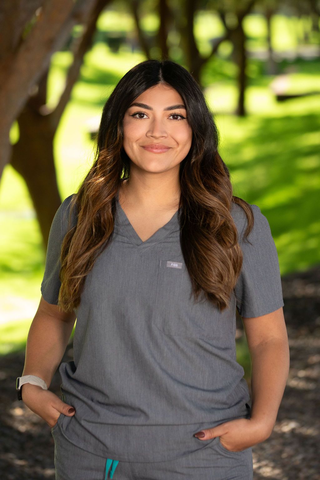 A woman in a gray scrub top is standing in front of trees with her hands in her pockets.
