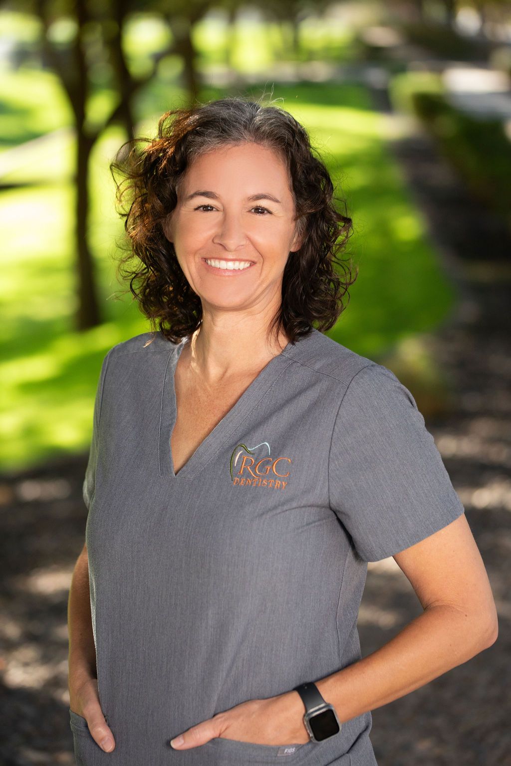 A woman in a scrub top is smiling and standing in front of a park