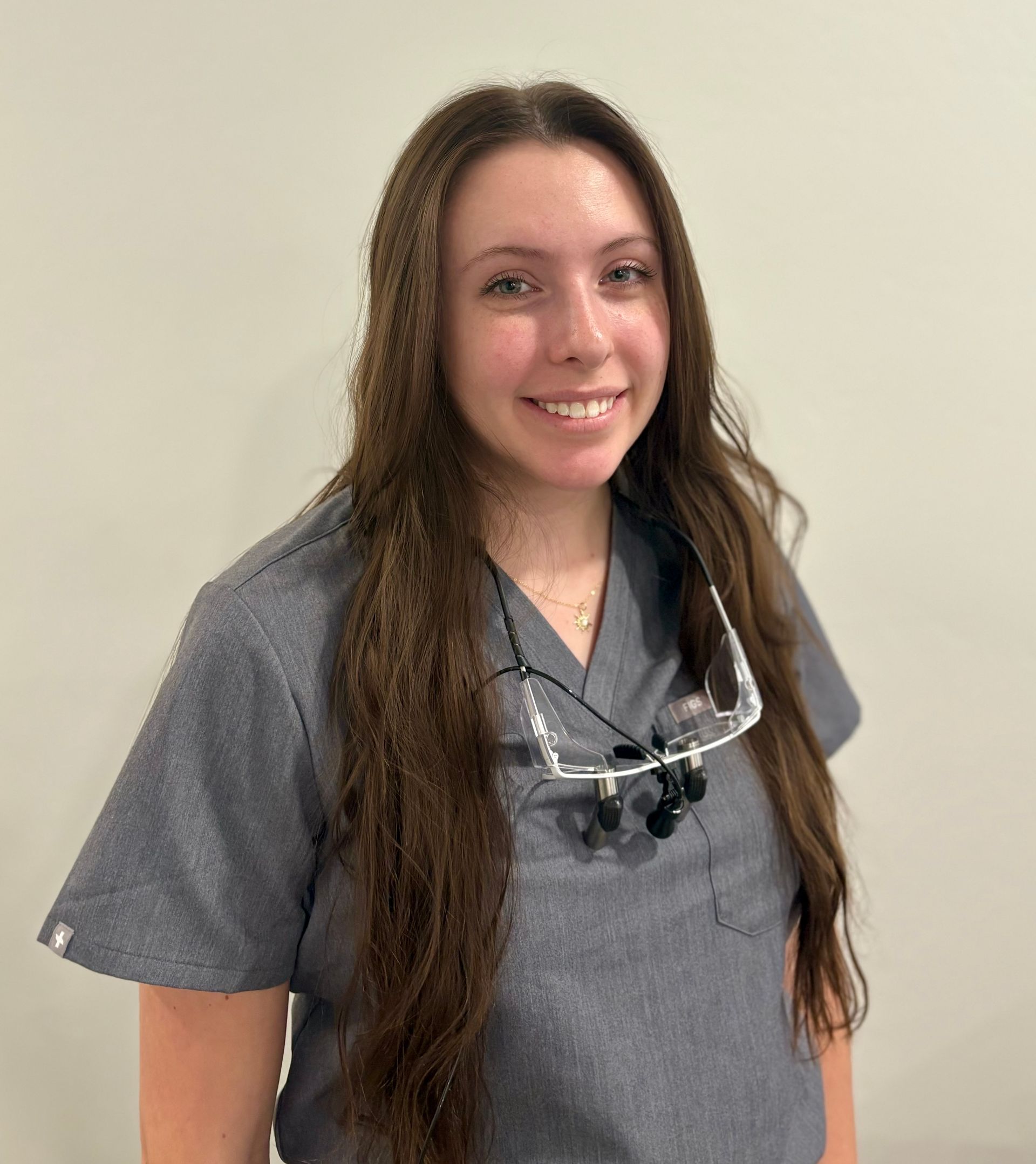 A woman in a gray scrub top is standing in front of trees with her hands in her pockets.