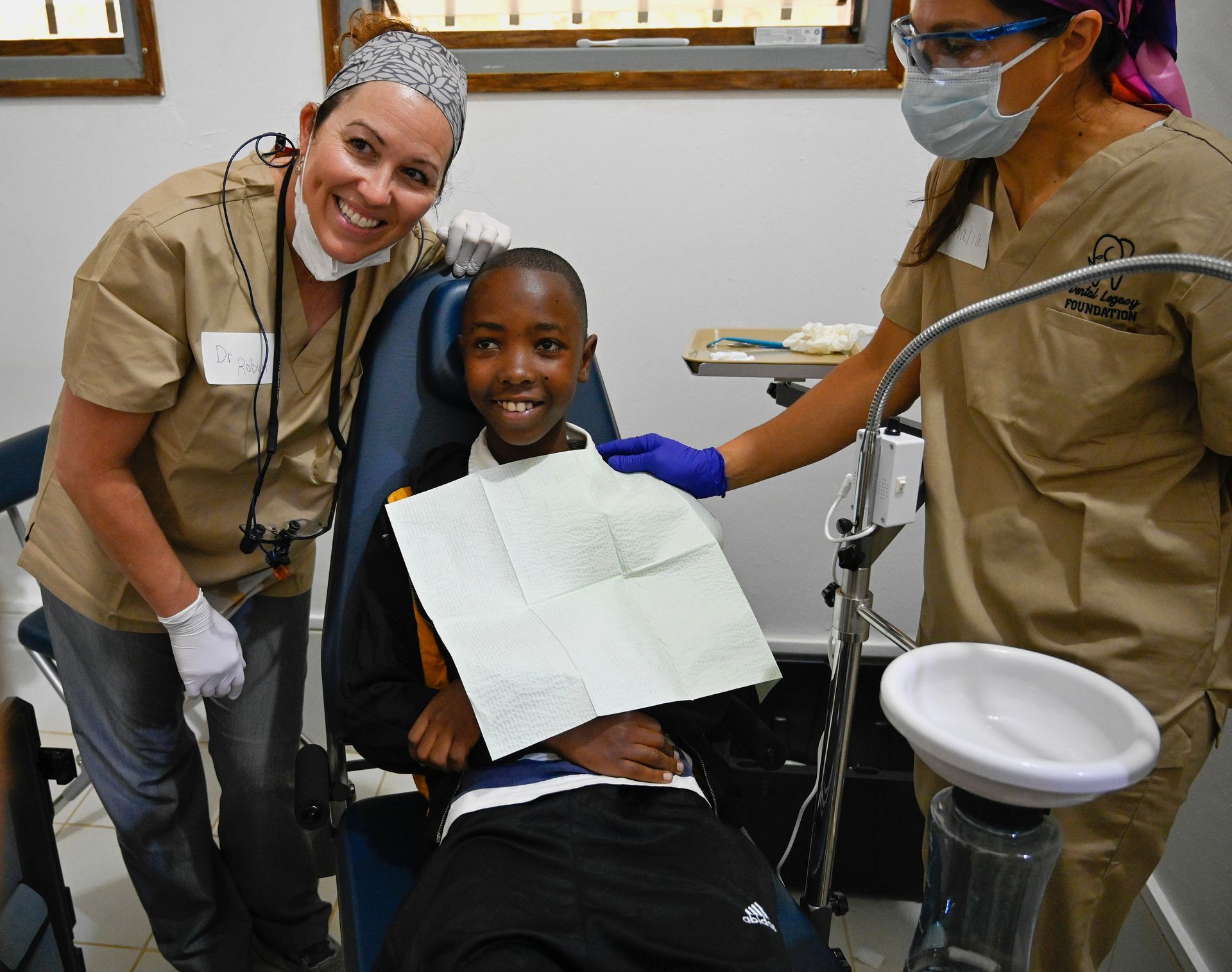 A young boy is sitting in a dental chair with two female dentists standing around him