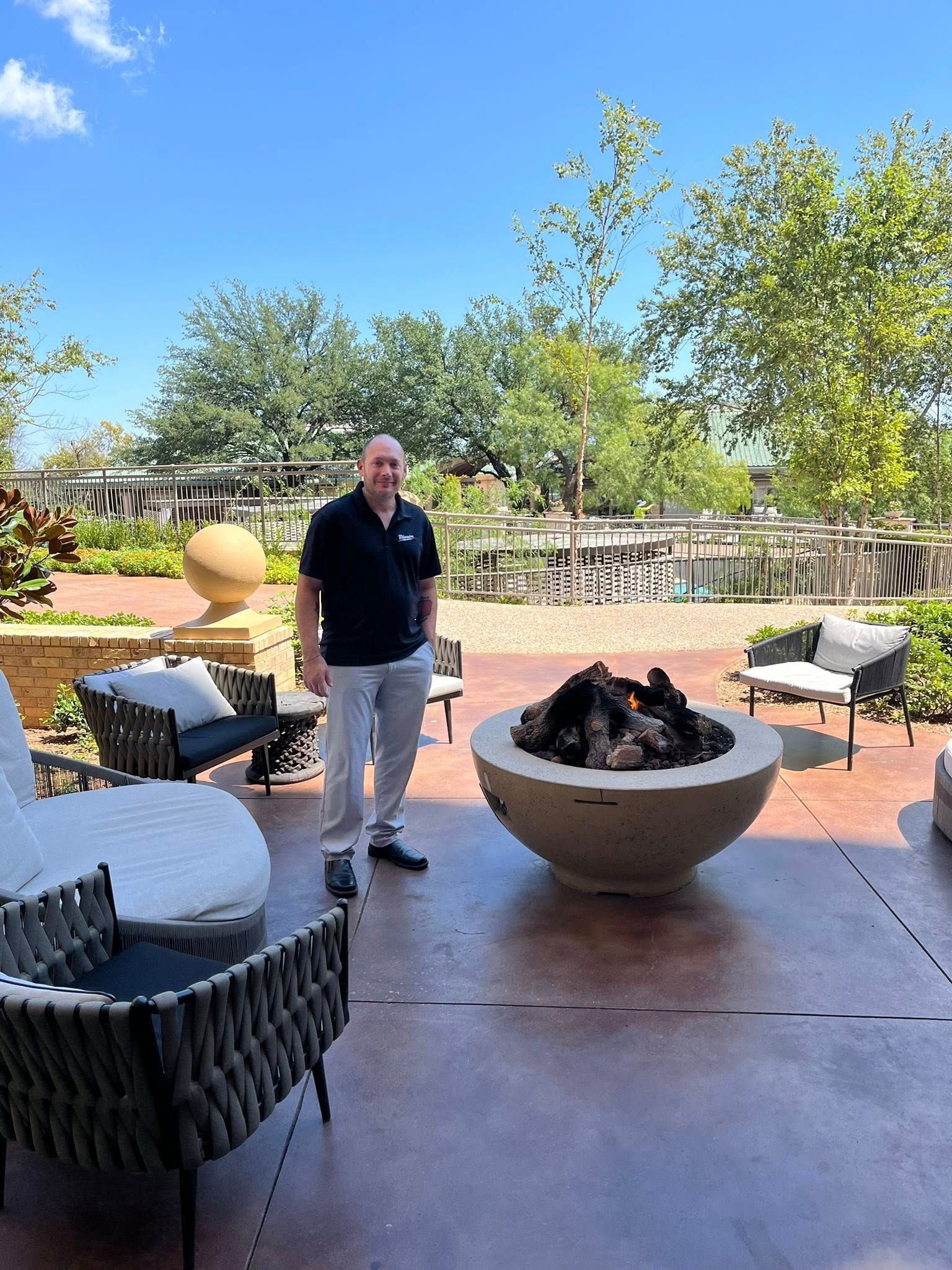 Man standing near a modern outdoor fire pit and patio furniture on a sunny day.