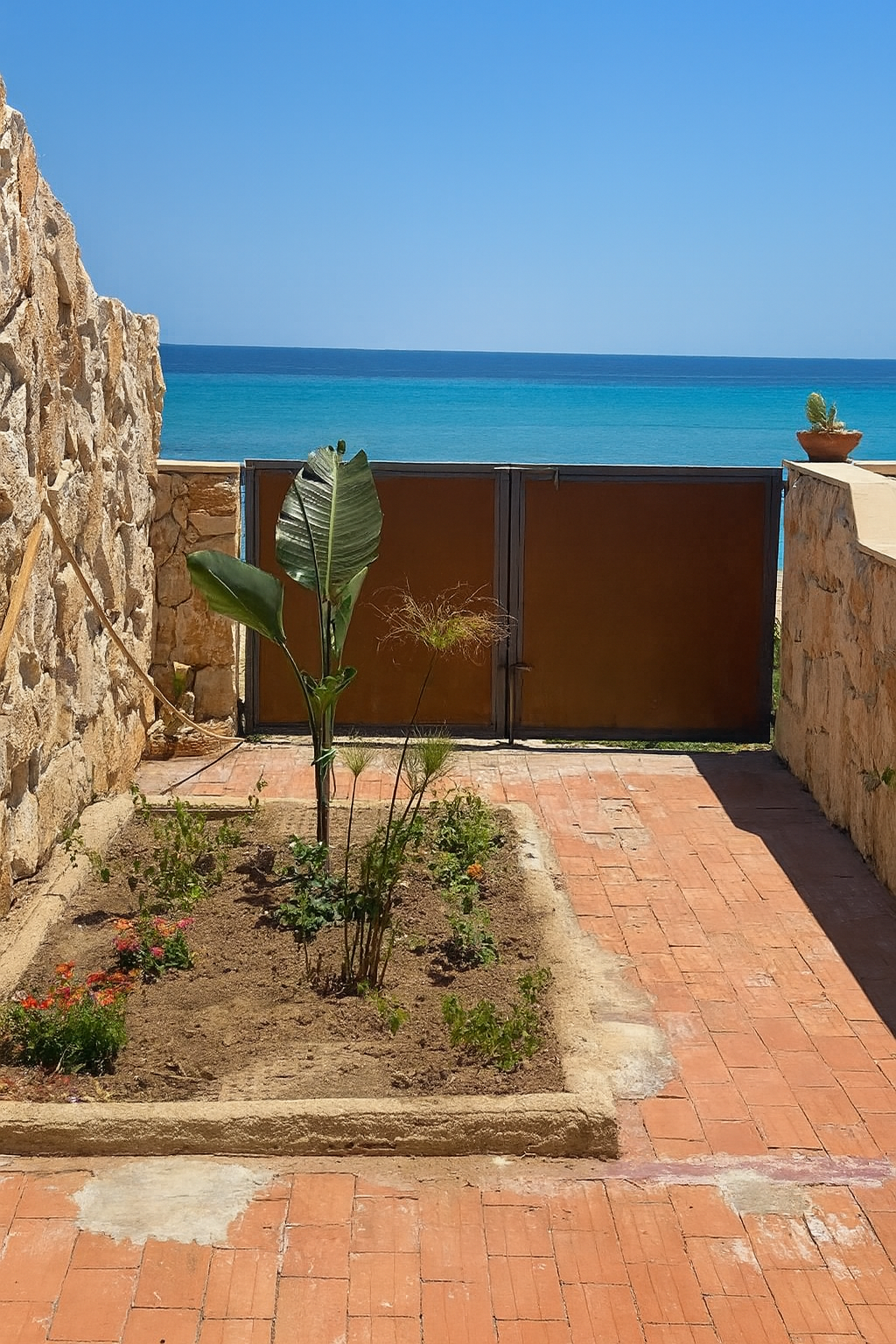 Giardino con piante e cancello che conduce all'oceano; muri in pietra incorniciano la vista dell'acqua blu e del cielo.