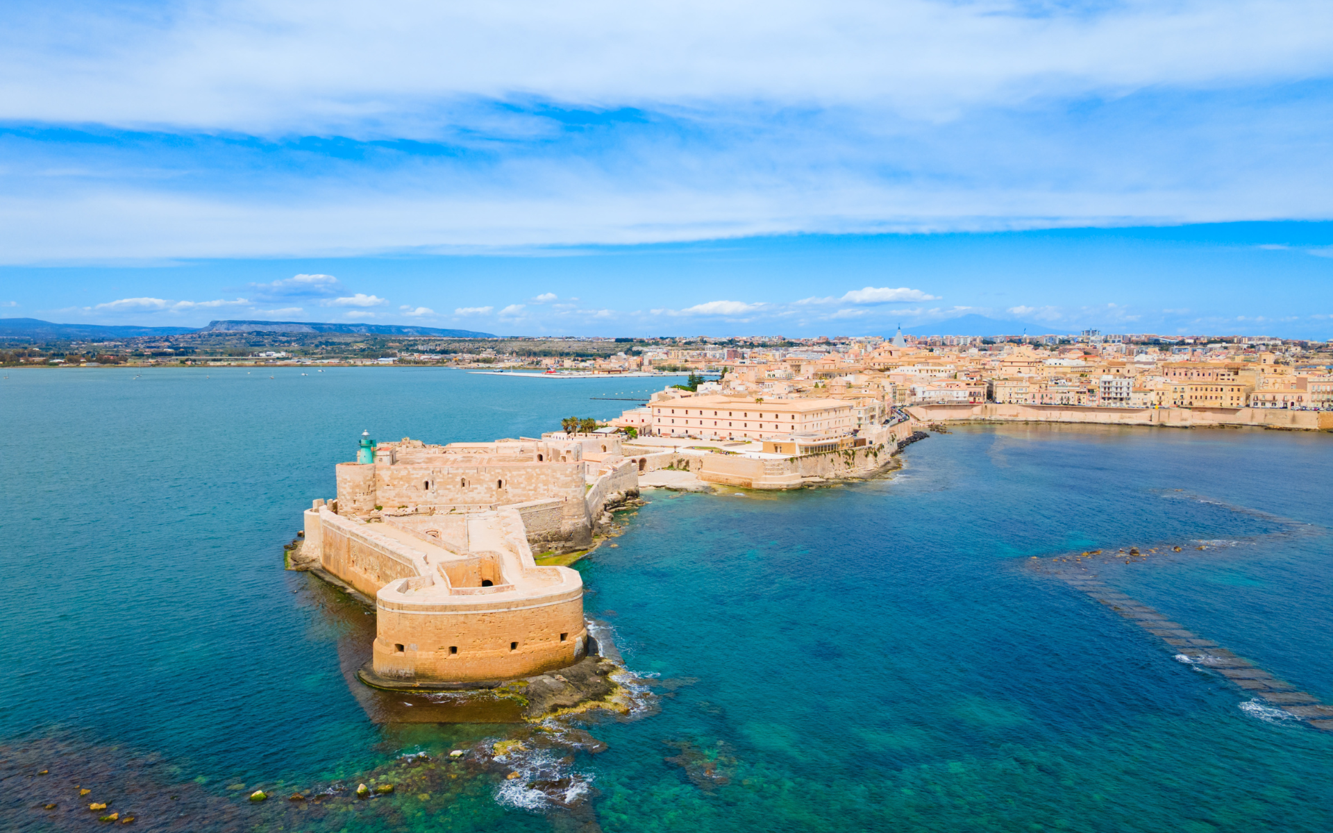 Vista aerea della costa di Siracusa, Italia, con un antico forte e il paesaggio urbano sull'acqua sotto un cielo azzurro.