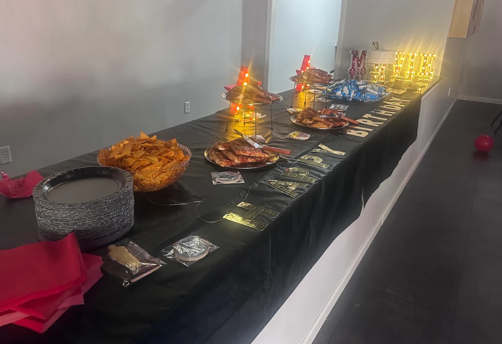 Long buffet table with assorted dishes, trays, and drinks on black tablecloth in a hallway