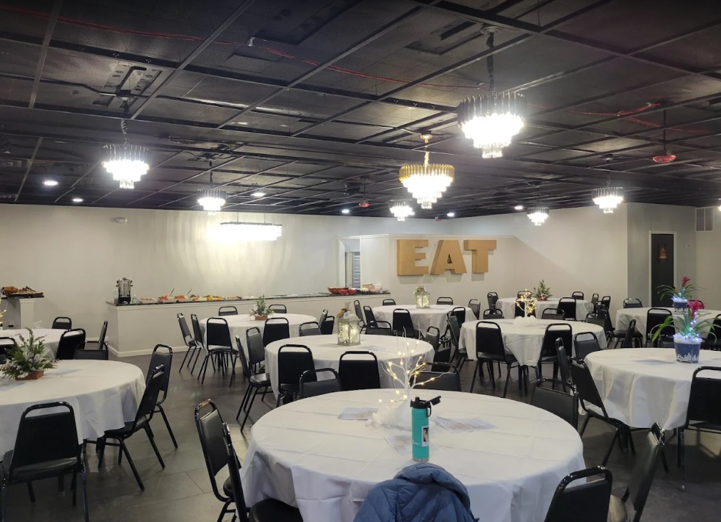 Large banquet hall with round tables and black chairs, wall letters spelling “EAT,” and hanging lights