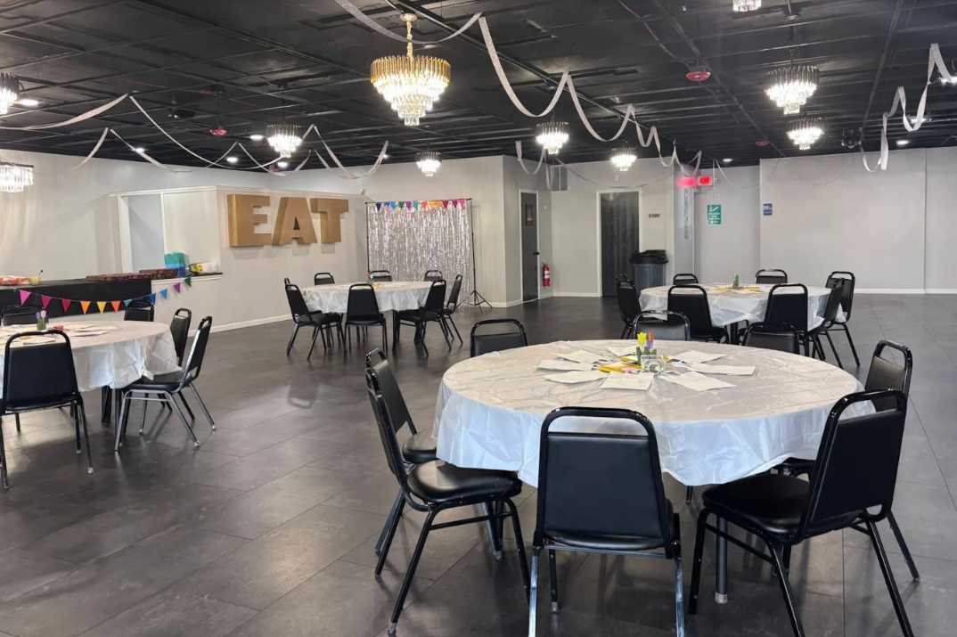 Banquet hall with round tables, black chairs, and “EAT” banner under hanging chandeliers