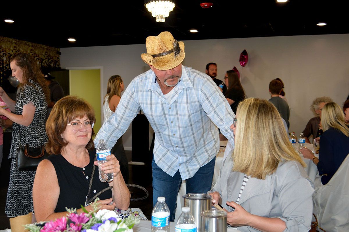 People chatting at a table as a man in a straw hat leans in at a lively indoor gathering.