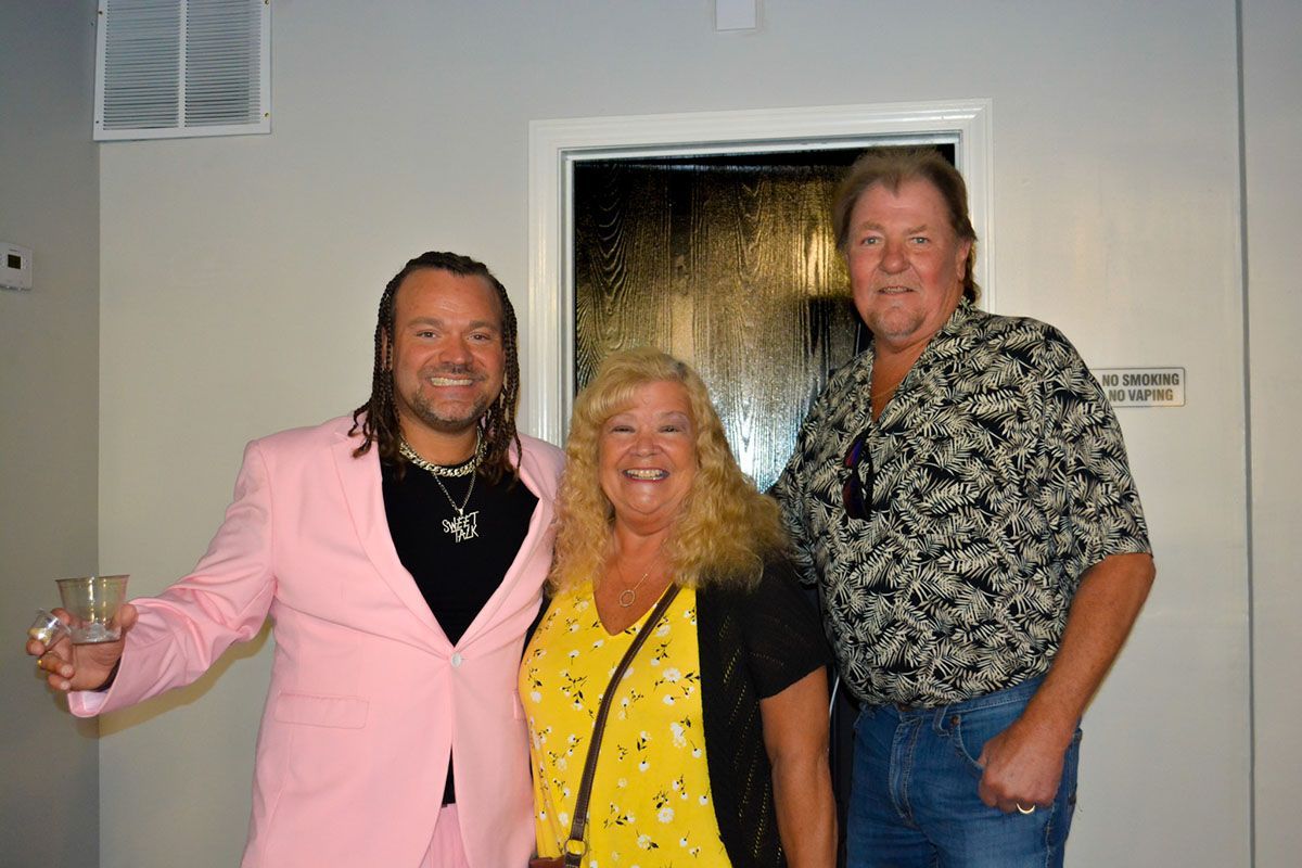 Three people posing indoors, smiling; man in pink suit holding a drink beside two others.