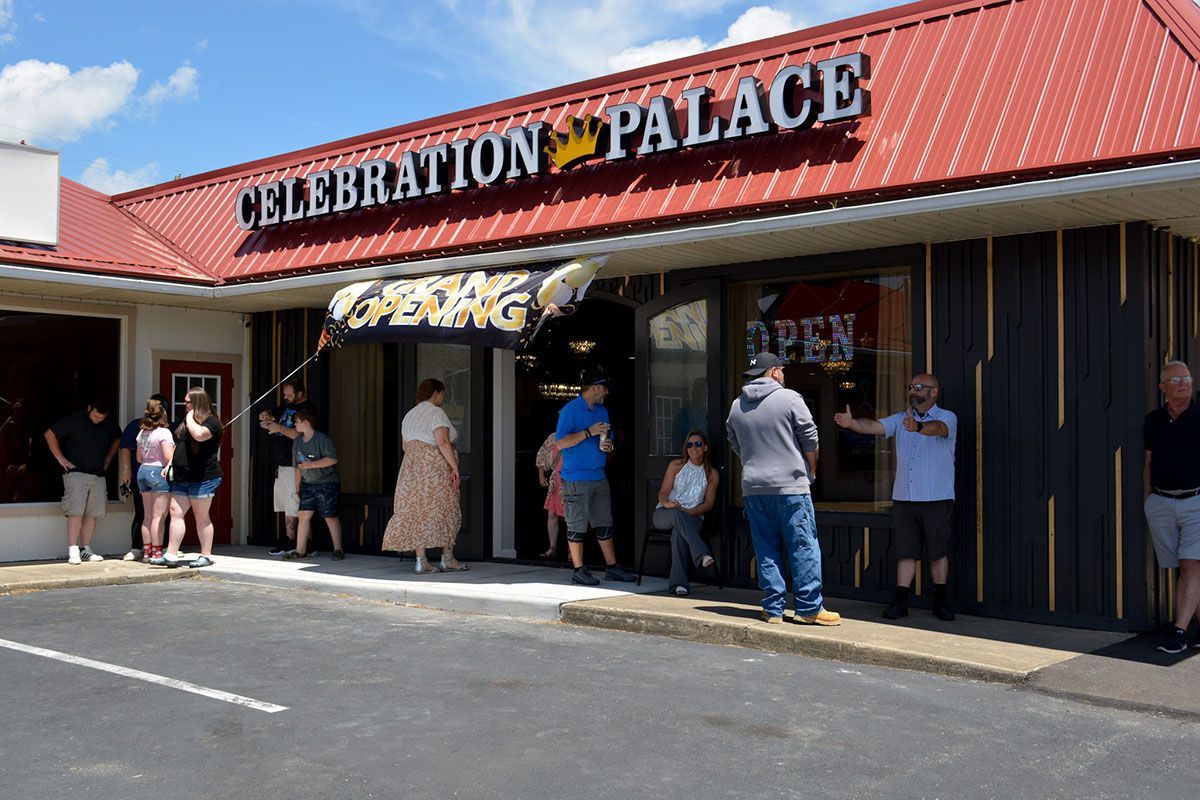 People standing outside Celebration Palace storefront under a red awning on a sunny day
