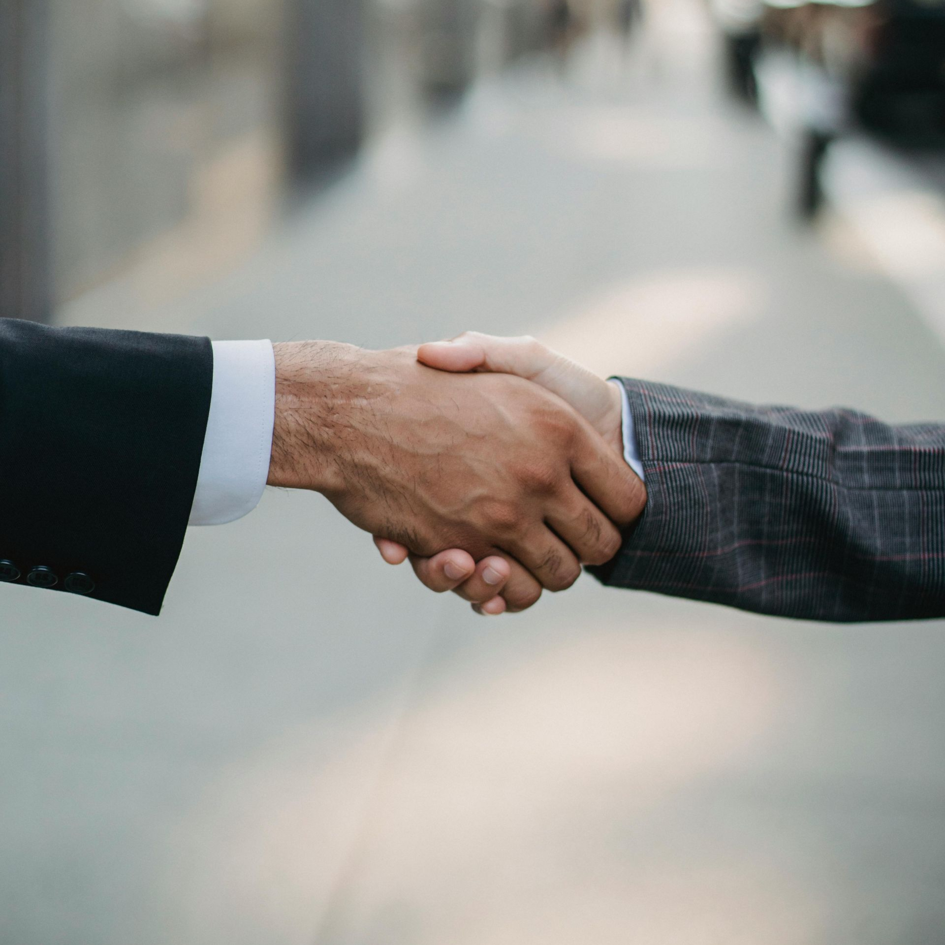 Two men in suits shaking hands, outdoors, blurred background.