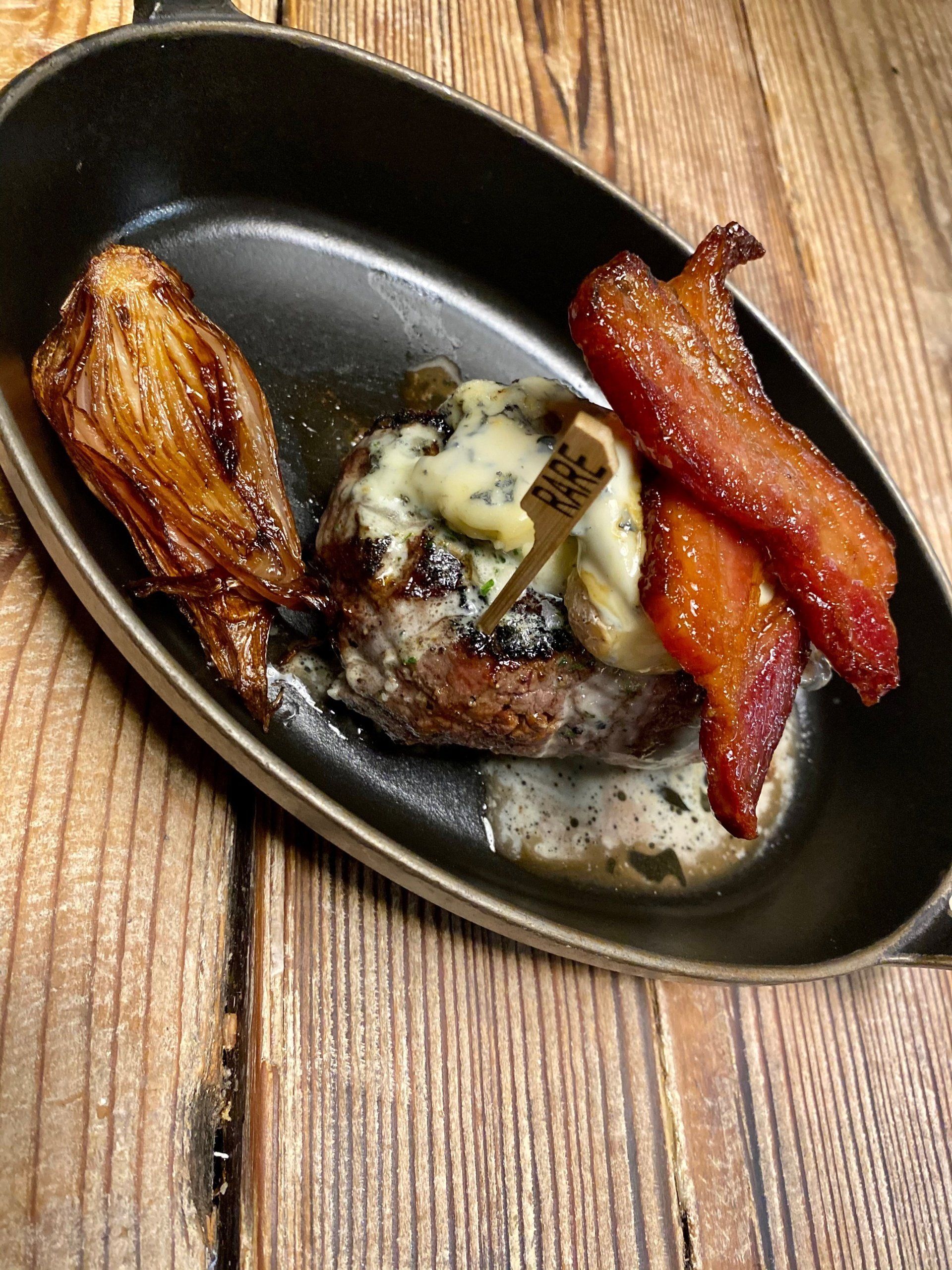 a plate of food with a steak and bacon on a wooden table