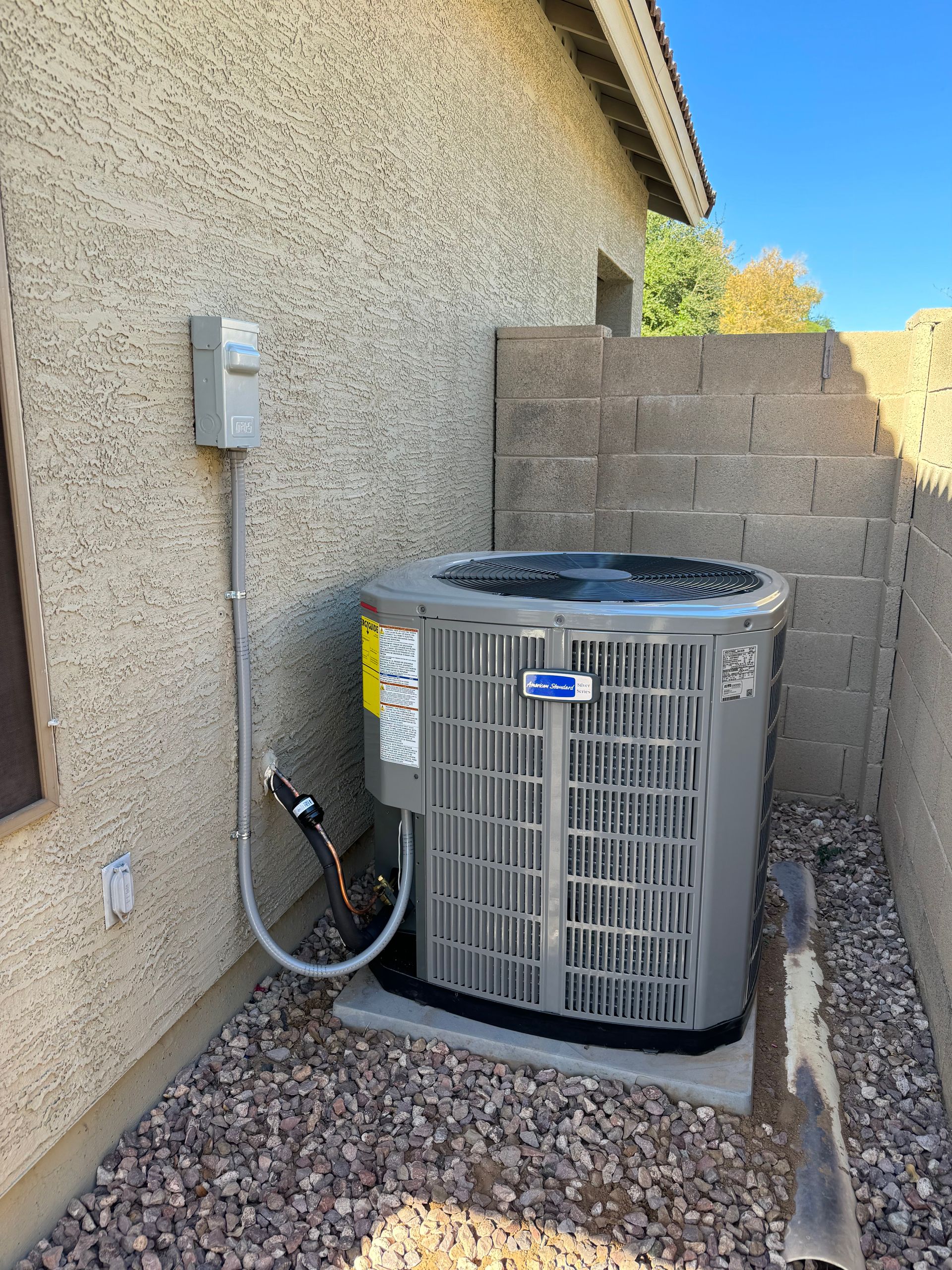 Outdoor HVAC unit beside a stucco house wall and cinderblock fence in a gravel yard.
