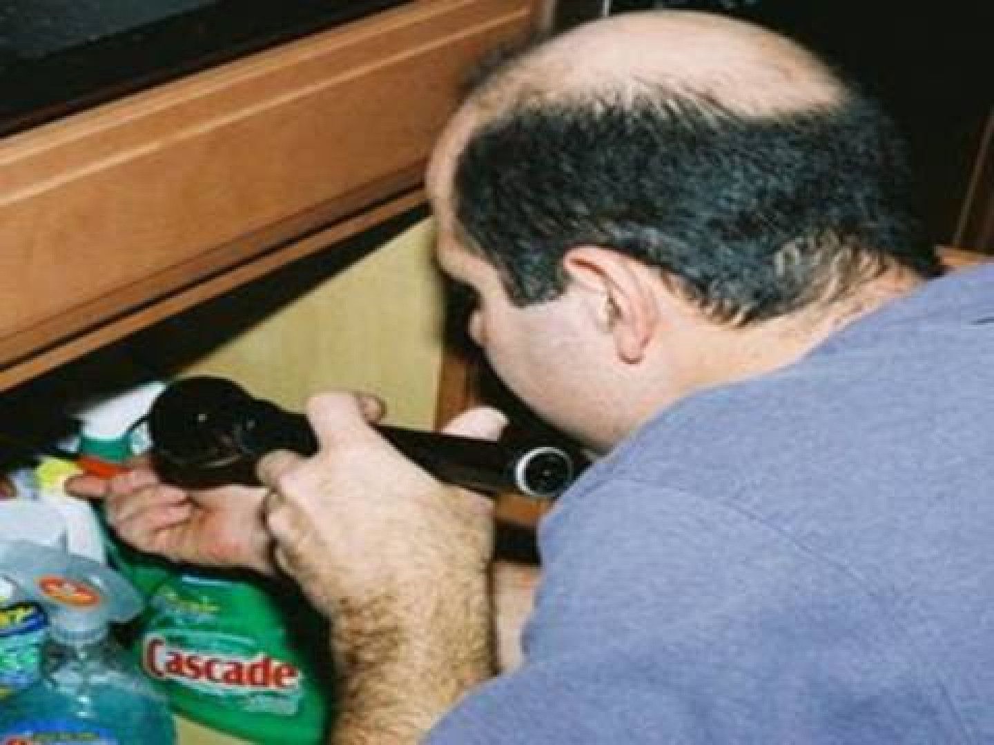 Man with balding head examines something under a cabinet with a flashlight.