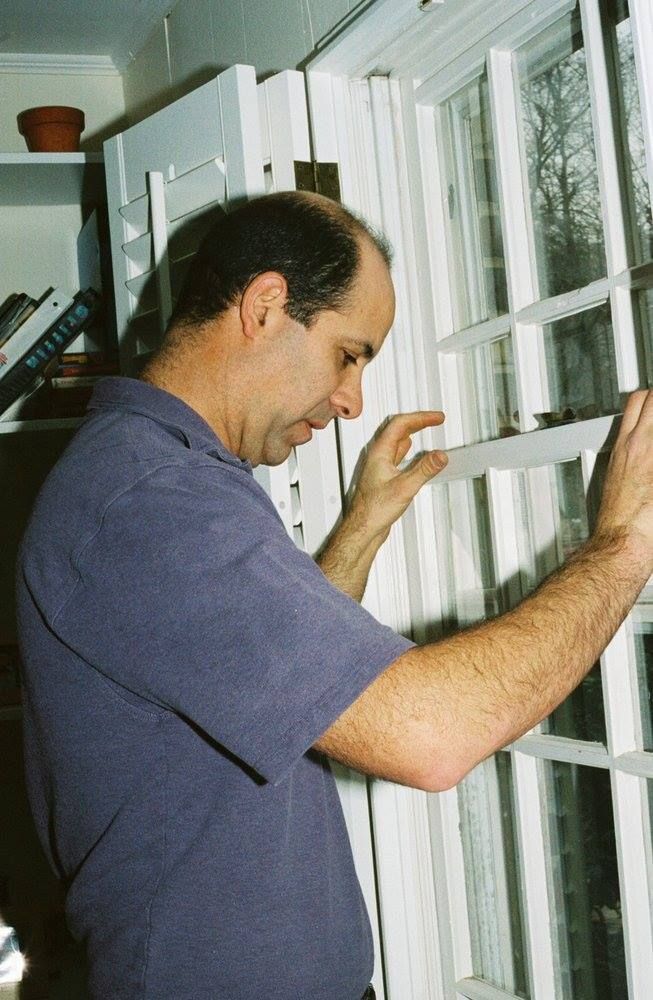 Man inspecting a white window, indoors. He wears a blue shirt and is focused on the window.