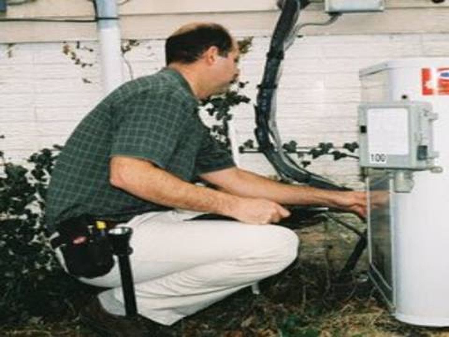 Man inspecting a gas meter near a building; he kneels with a tool belt.