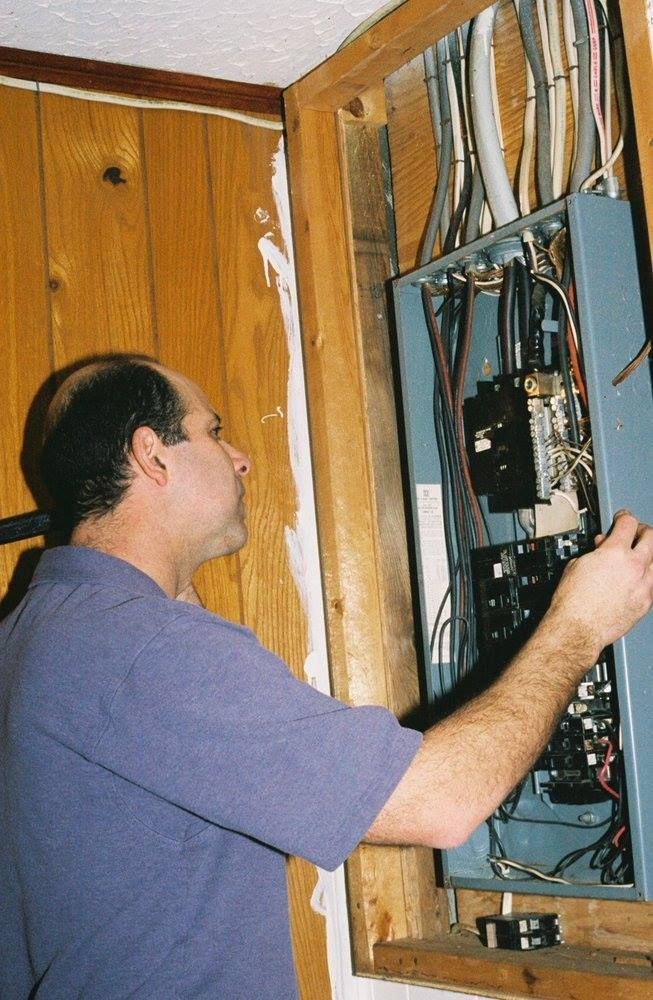 A person inspecting an electrical panel in a wooden wall. Wires and breakers are visible.