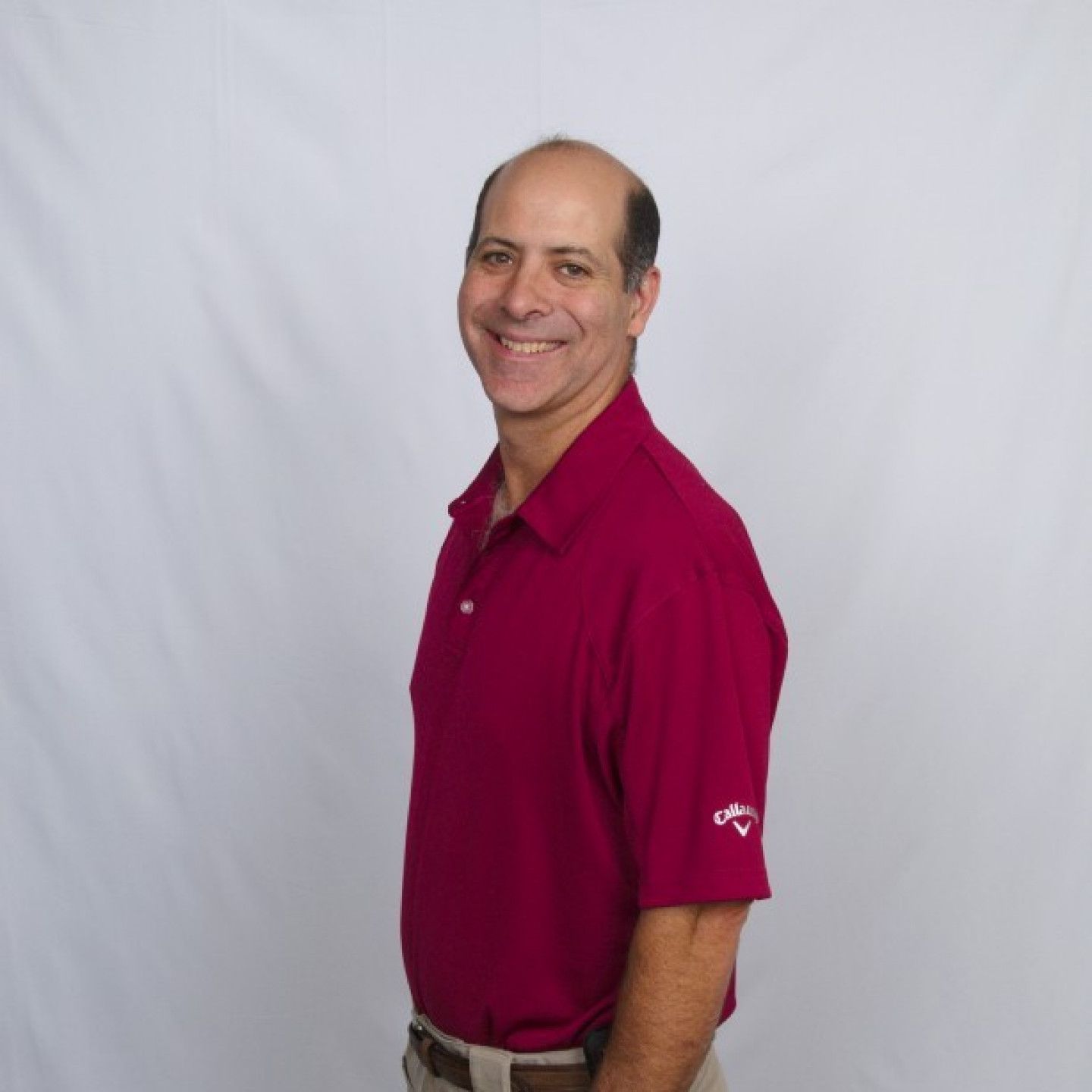 Man in maroon polo shirt smiles, stands sideways against a white backdrop.