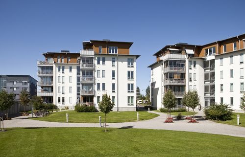 Apartment buildings with white facades, balconies, and brown roofs on a sunny day. Green lawn in the foreground.