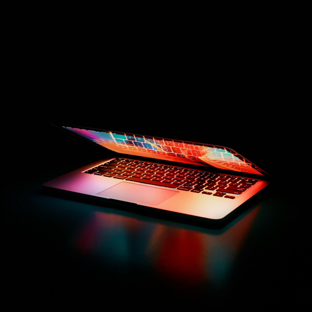Laptop mit geöffnetem Bildschirm, der in buntem Licht erstrahlt, vor dunklem Hintergrund.
