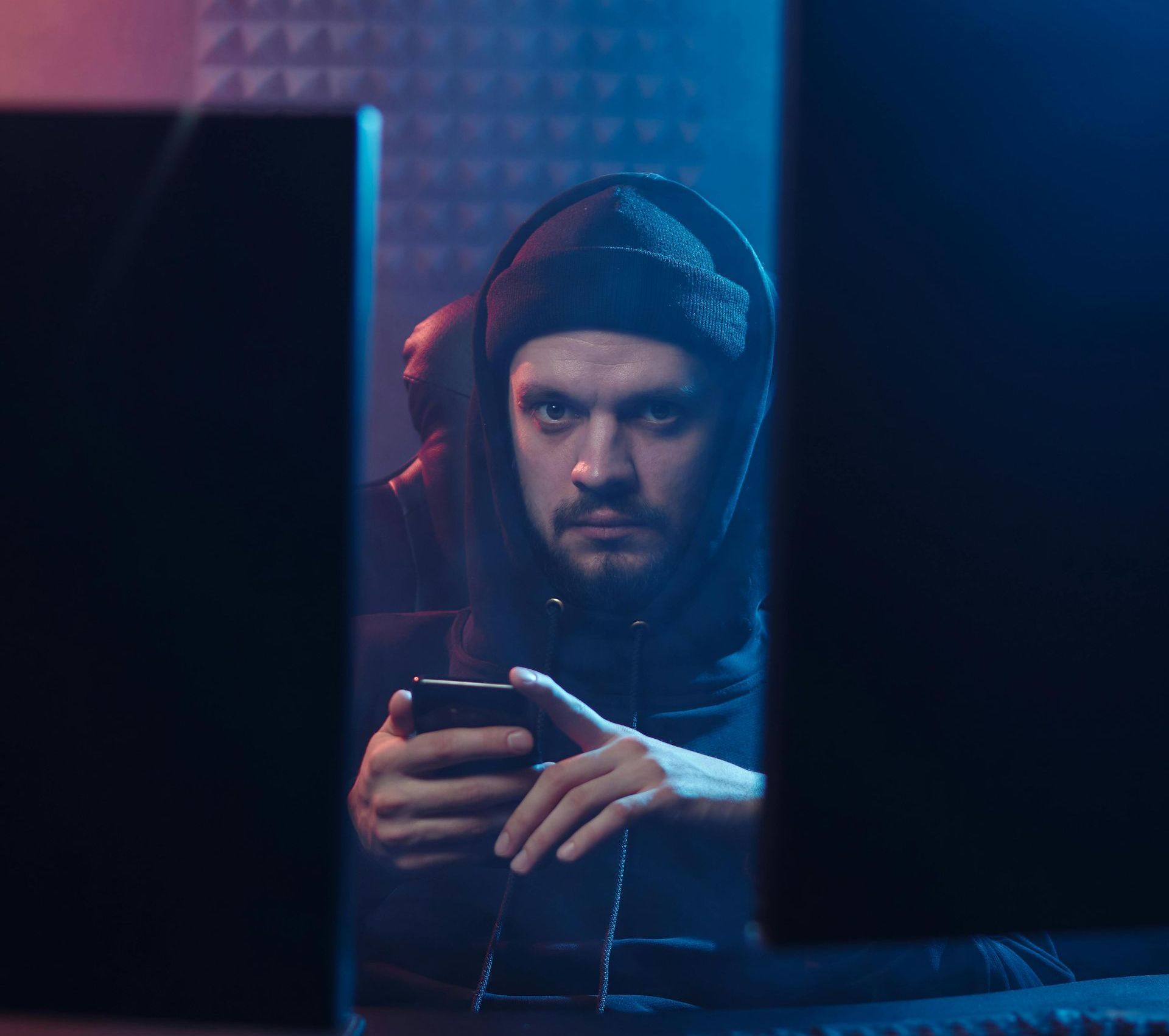 Ein Mann in einem Kapuzenpullover blickt konzentriert auf ein Handy, das von zwei Computerbildschirmen flankiert wird, unter blauem und rotem Licht.
