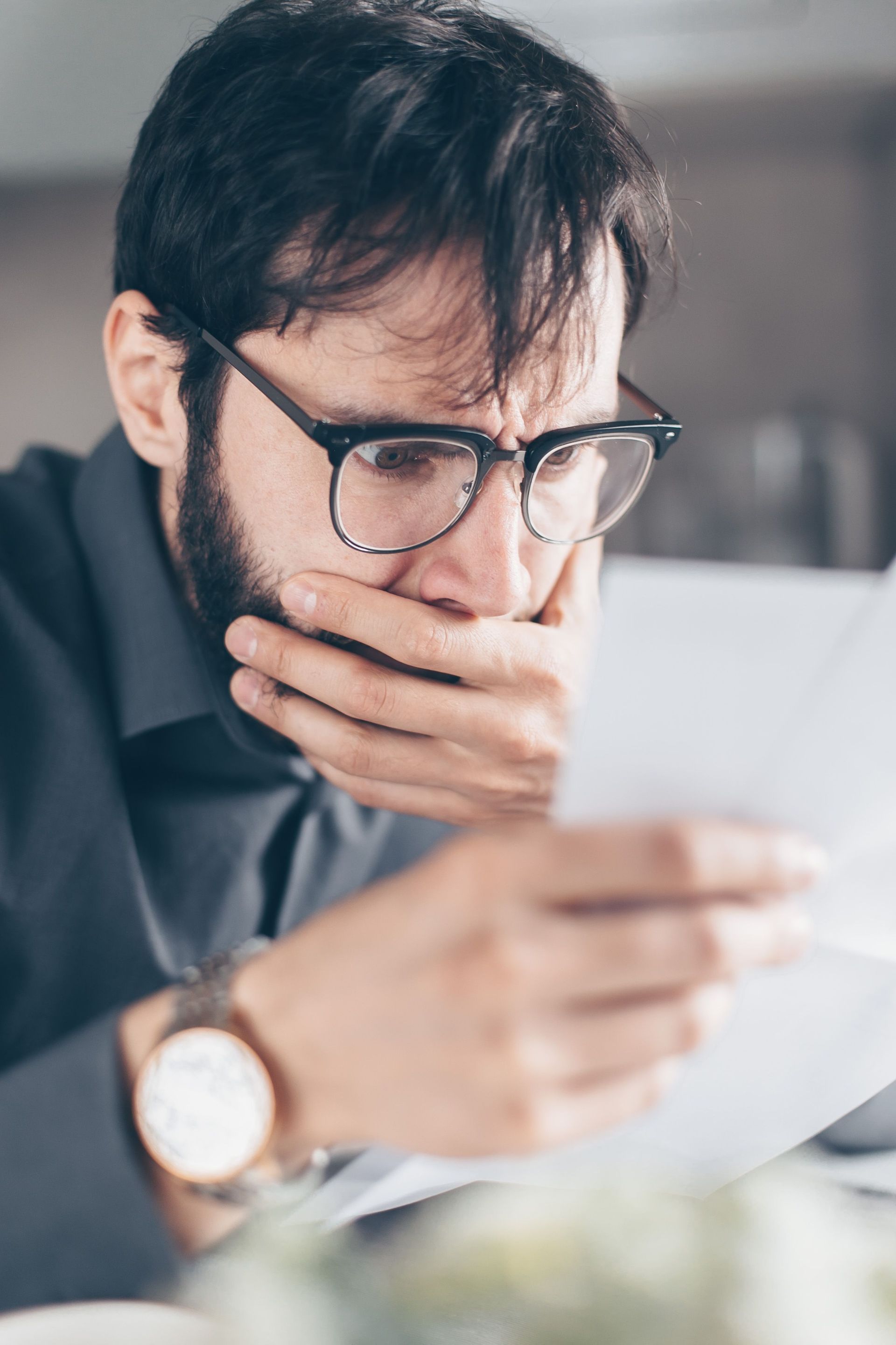 Man in glasses reading a document with a hand over his mouth, appearing concerned at a desk