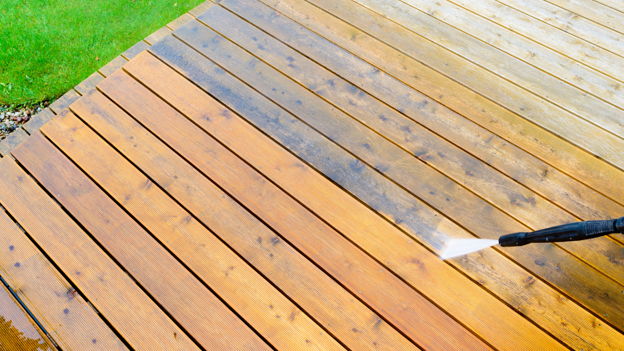 Pressure washing a wooden deck with a black hose, cleaning stained boards near green grass.