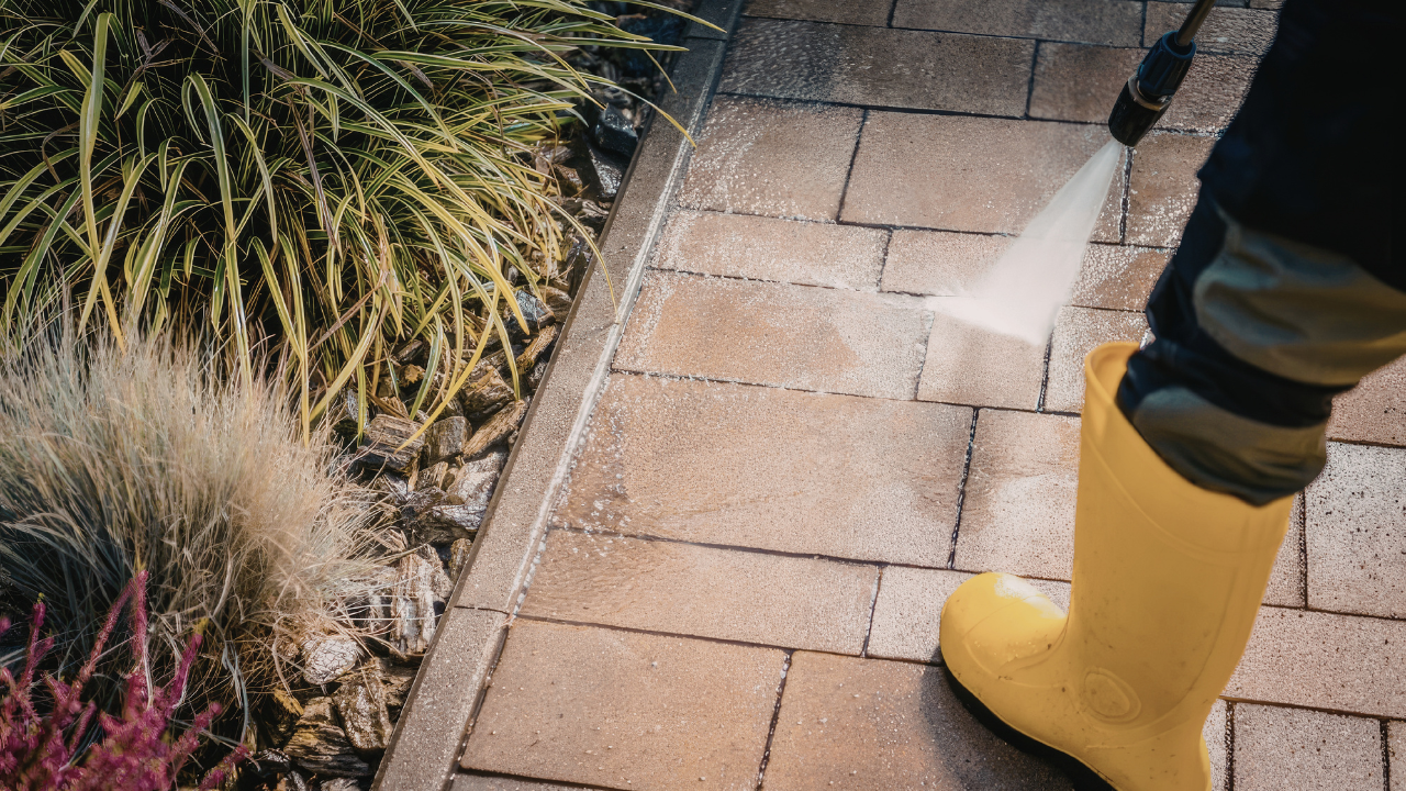 Person in yellow boots spraying a brick walkway beside garden plants