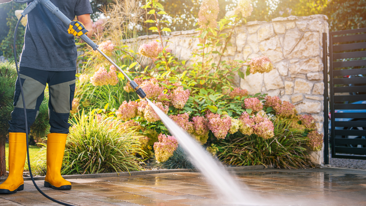 Person pressure-washing a wet driveway beside a flower bed and stone wall, wearing yellow rain boots
