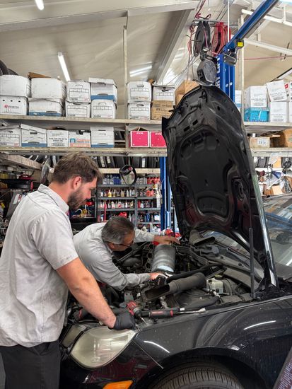 Two mechanics working on a car engine inside a cluttered auto repair shop. One wears gloves, examining parts. | Pacifica Auto Works