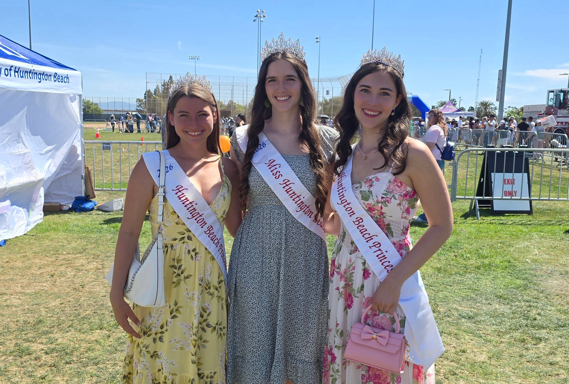 Three people wearing crowns and blue superhero capes pose for a photo in front of a comic book-themed backdrop outdoors.