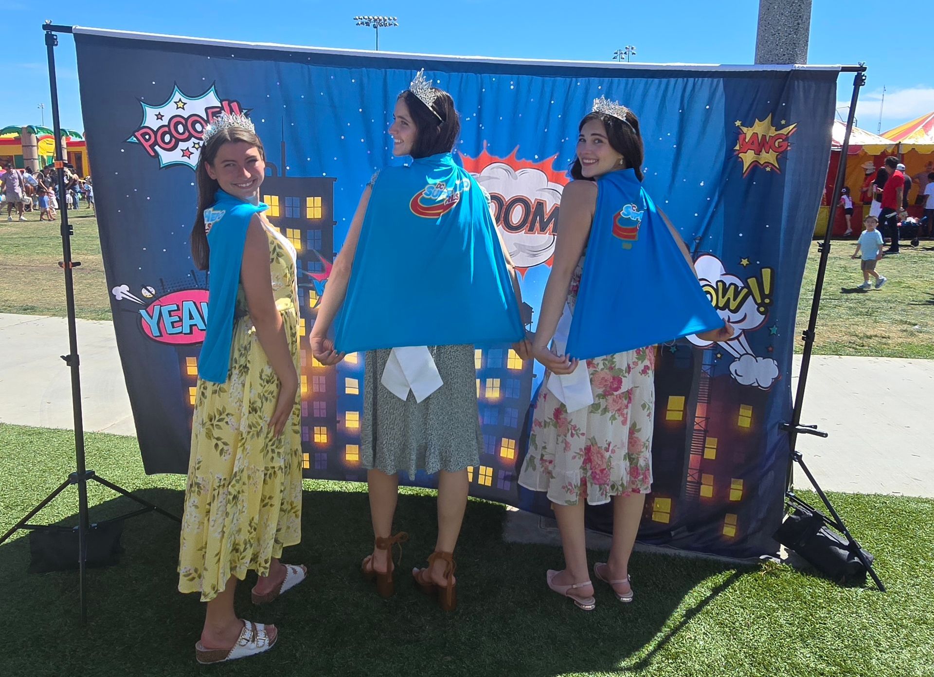 Three people wearing crowns and blue superhero capes pose for a photo in front of a comic book-themed backdrop outdoors.