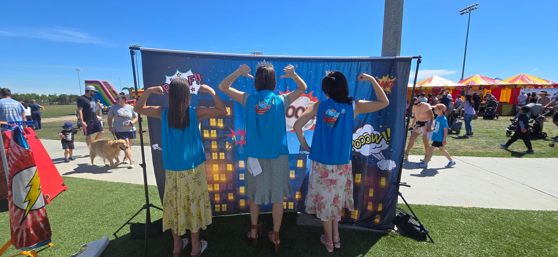 Three people wearing crowns and sashes stand before a comic-themed backdrop with their arms raised in celebration.