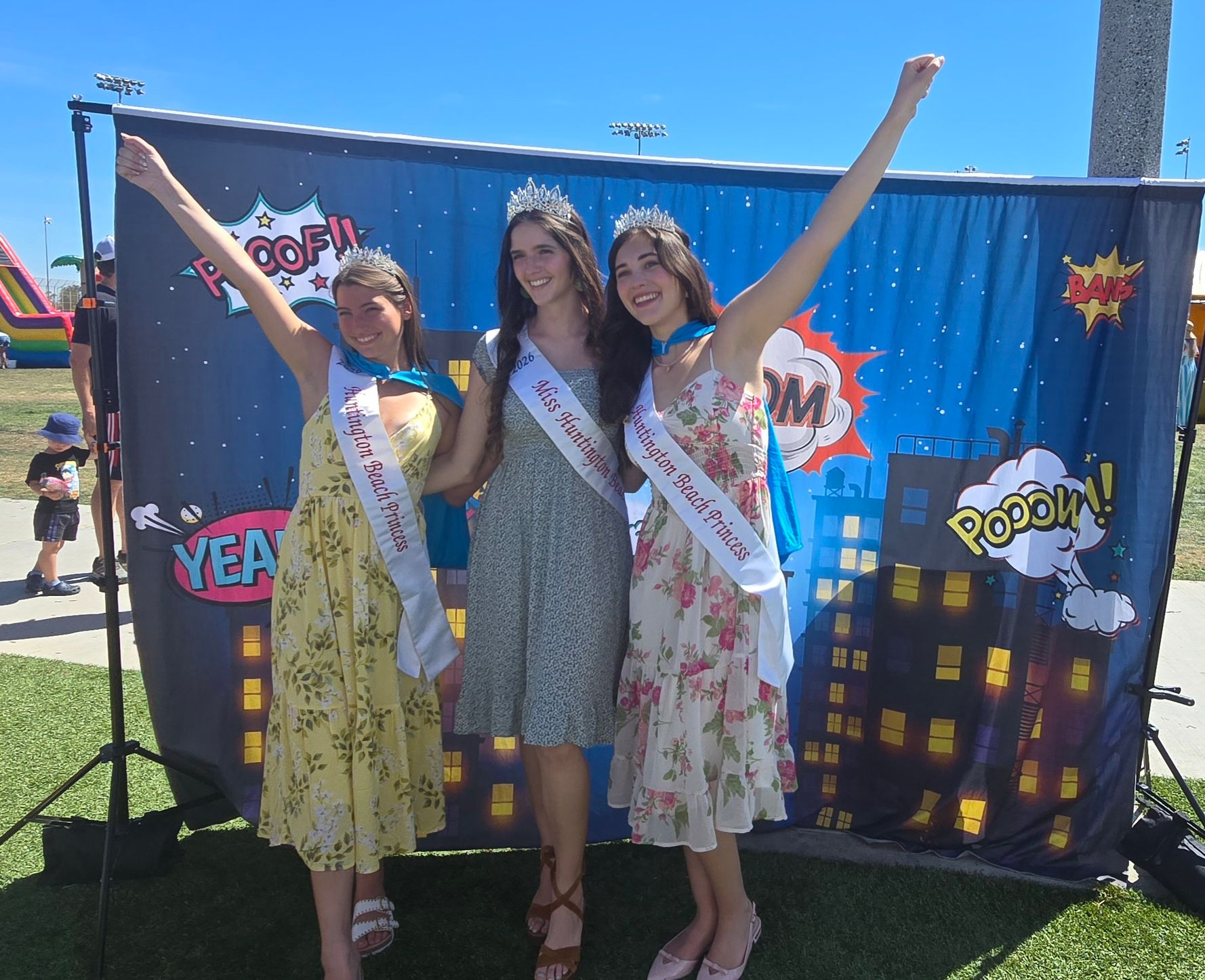 Three people wearing sashes and tiaras pose with arms raised in front of a comic book-themed backdrop at an outdoor event.