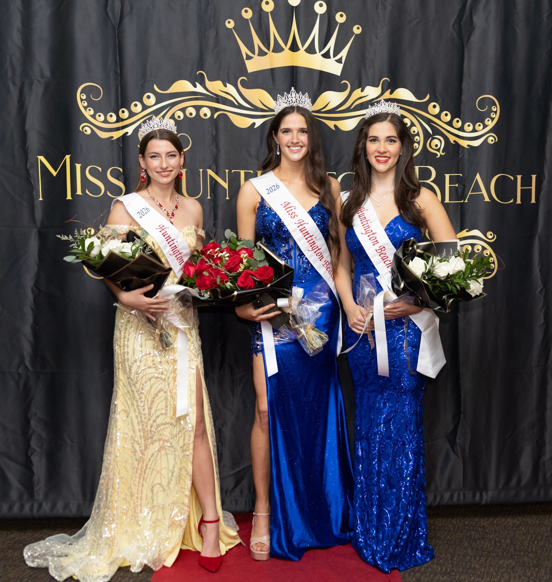 Three people wearing evening gowns and tiaras hold bouquets in front of a “Miss Huntington Beach” banner.