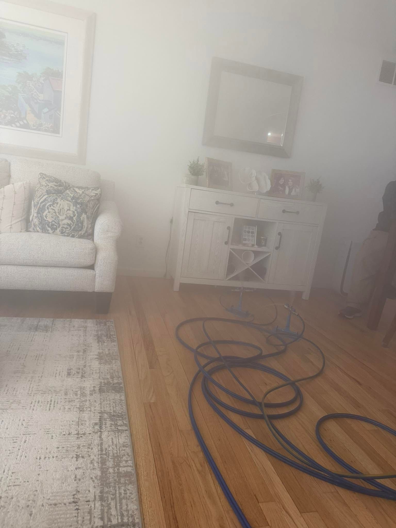 Smoke-filled living room with armchair, side table, mirror, and coiled blue hose on hardwood floor