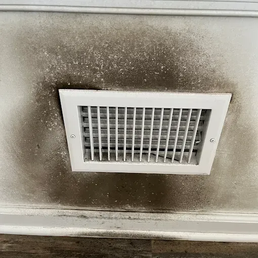 Wall vent with dark soot or mold stains above a worn wood floor under a window.