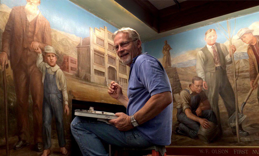 image of an old white man sitting in front of a historic mural with a brush in hand and a palette of art conservation pigment on his lap