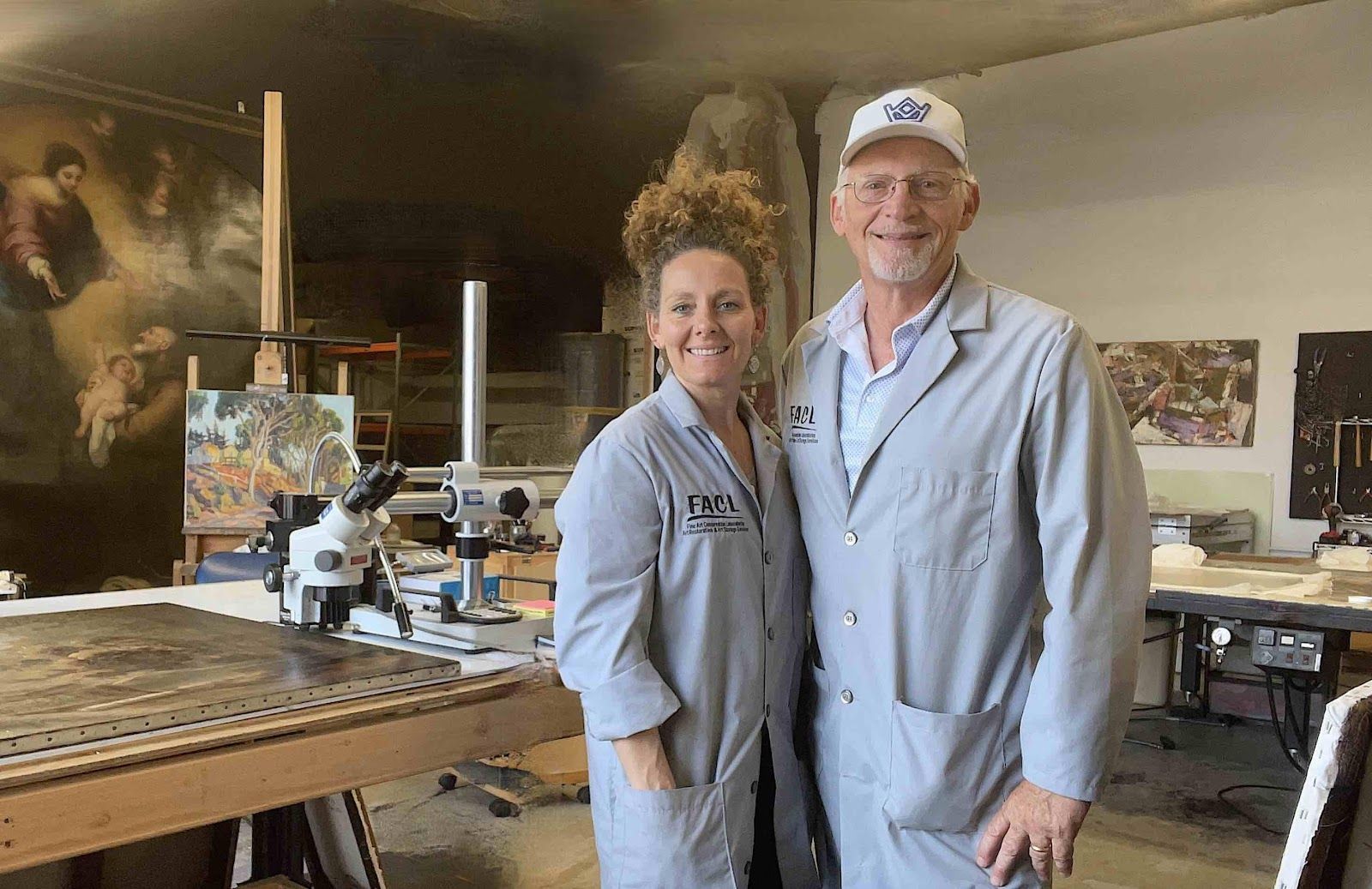 an old white man and a middle-aged white woman wearing lab coats in an art conservation lab stand side by side smiling