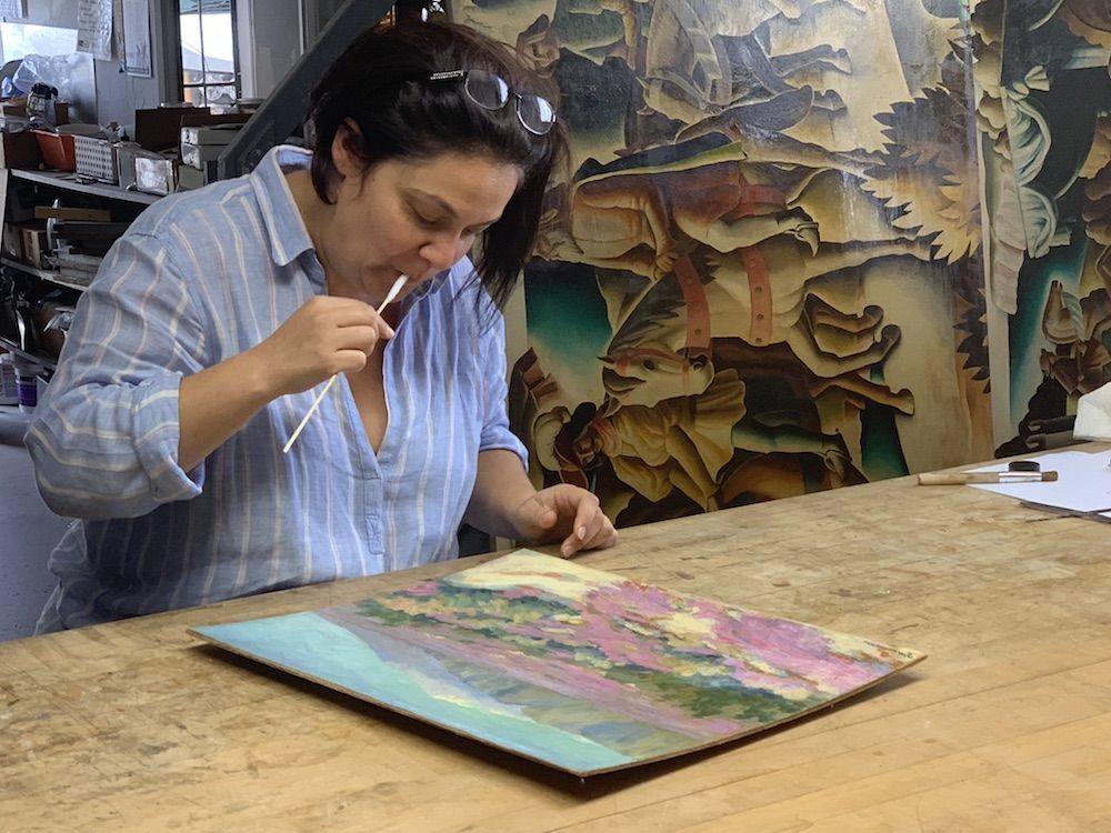 A female art conservator applies saliva to a cotton swab to prepare for cleaning a painting 