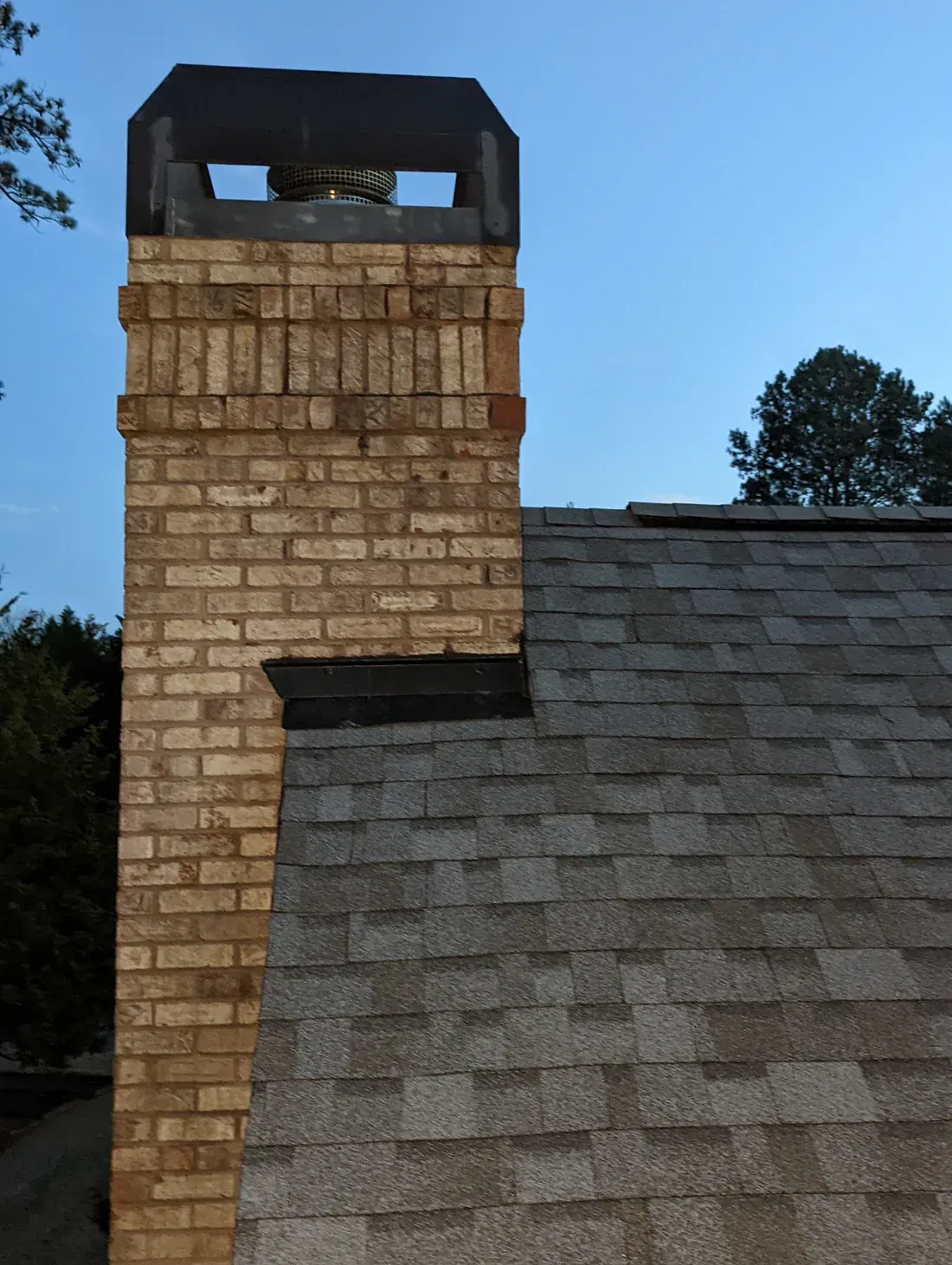 Brick chimney on a shingled roof against a blue sky, topped with a black cap.