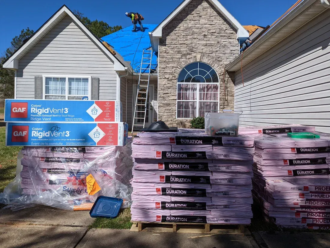 Roofing materials stacked in front of a house. A worker is on the roof with a blue tarp.