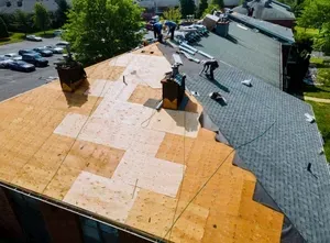 Workers installing roofing on a building, partially completed. Wood panels and asphalt shingles visible.