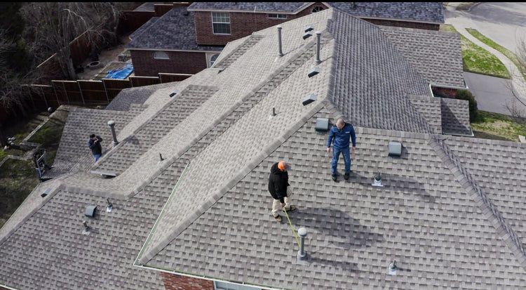 Two people stand on a gray shingled roof, inspecting it. Chimneys and vents are visible.