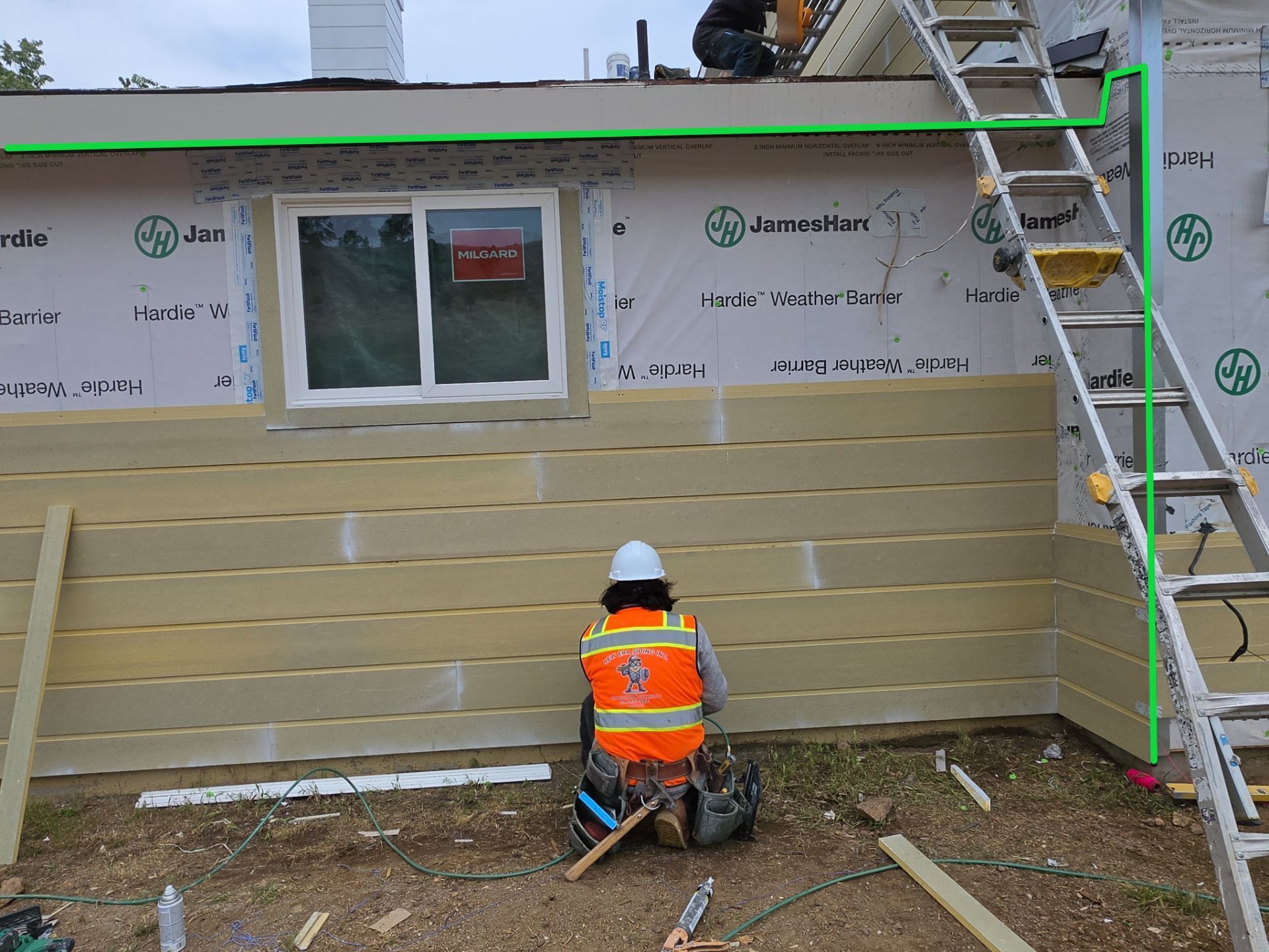 Worker in orange vest installing siding on a house exterior beneath a window, with a ladder nearby.