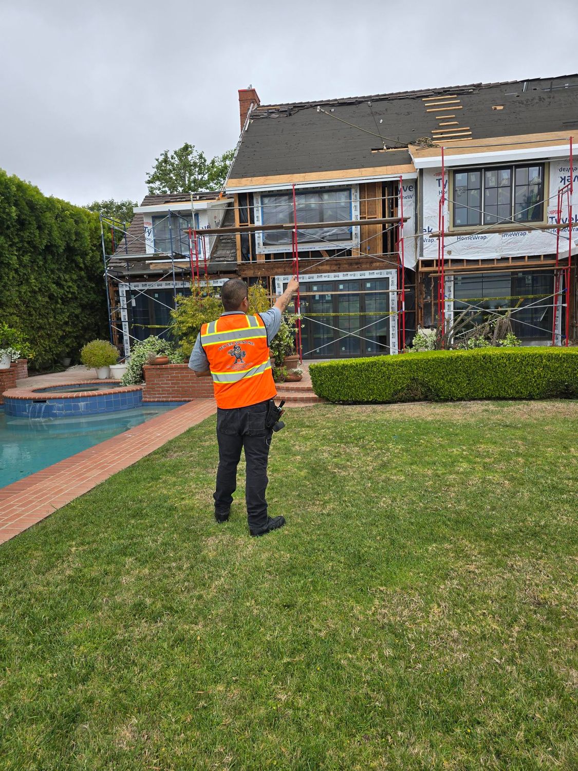 A person in a high-visibility vest stands in a yard and gestures toward a house undergoing exterior renovations.