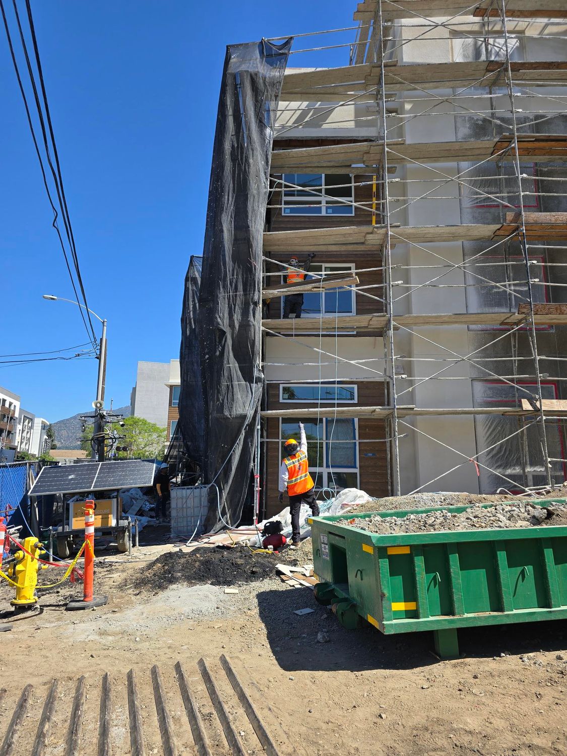 Construction workers on scaffolding in front of a building under renovation, with a large green dumpster in the foreground.
