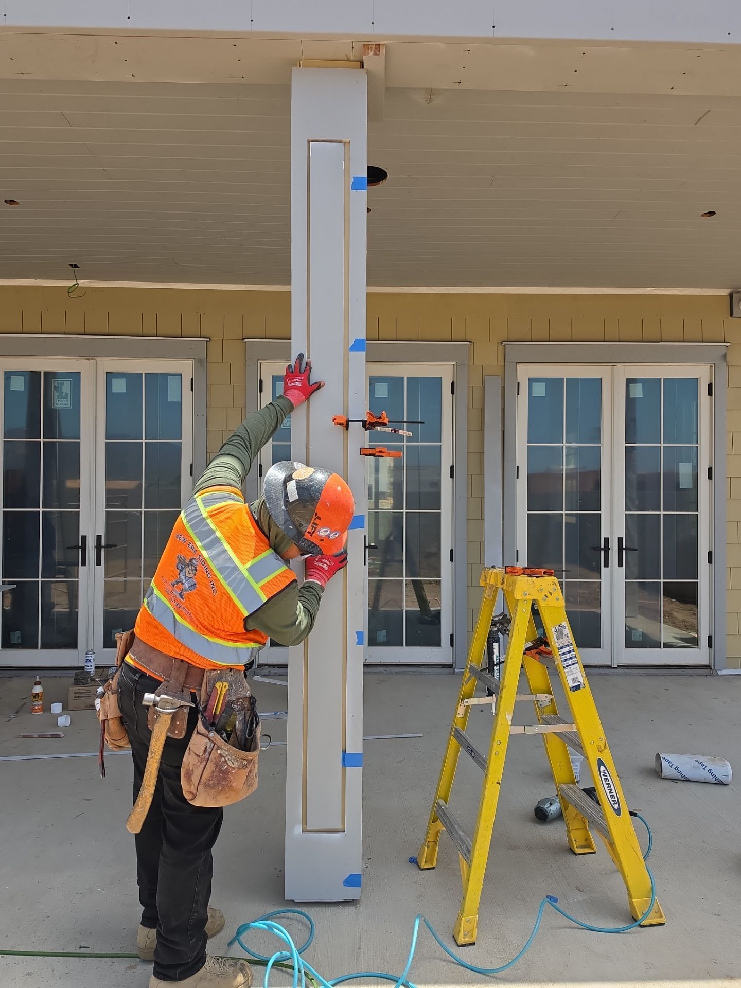 Worker in orange vest adjusting a tall column with a ladder outside a building.