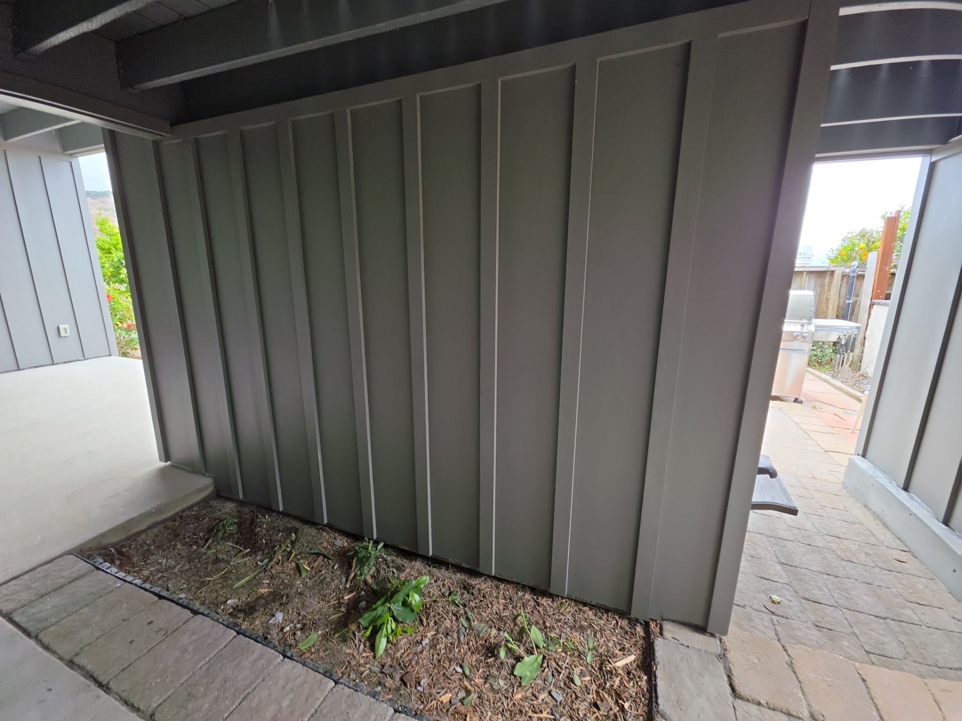 Metal shed wall beside a narrow dirt planter and concrete walkway under a covered patio
