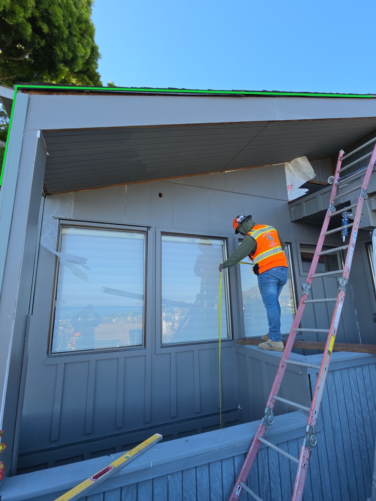 Worker in orange vest painting the exterior of a house from a ladder under the eaves.