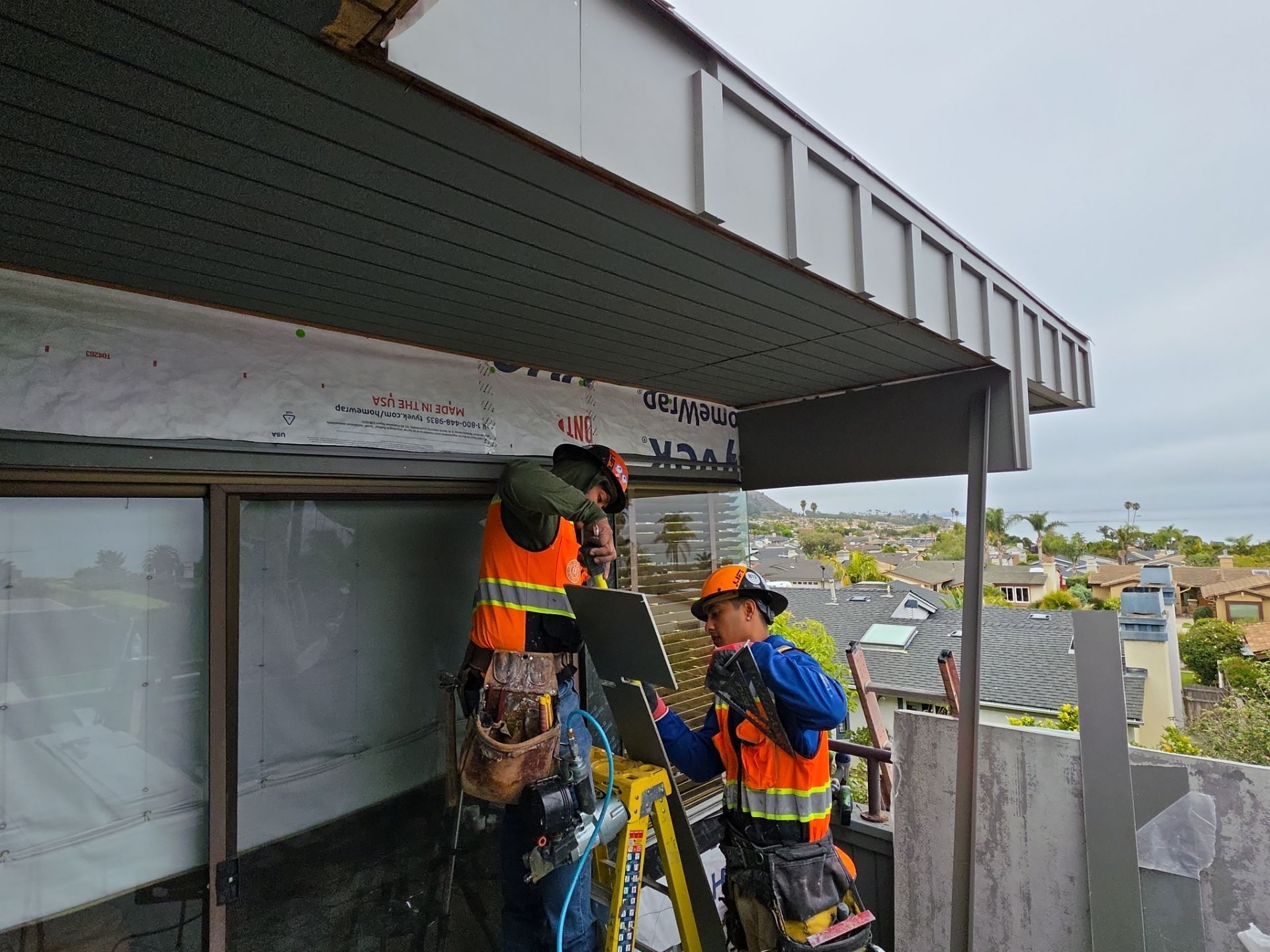 Two workers in safety gear repairing the underside of a roof on a building exterior
